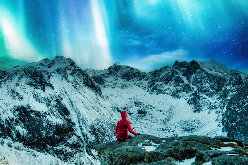 Rear view of mountaineer in red jacket sitting on the cliff with Aurora borealis over snowy mountain on Ryten in Lofoten Islands, Norway