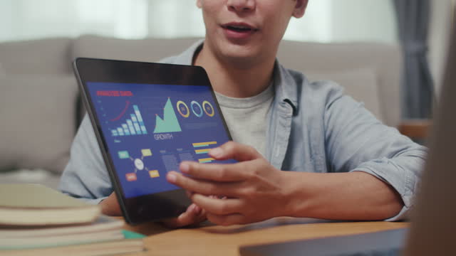 Close-up shot of a Young man sitting in cozy living room and presenting business stock market, trading, infographic, and charts on a tablet with colleague via video conference from home.