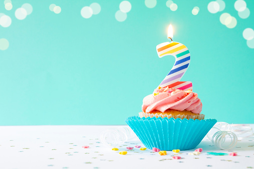 Vanilla buttercream cupcake with pink icing, sprinkles and 2nd birthday candle, on a light blue background.