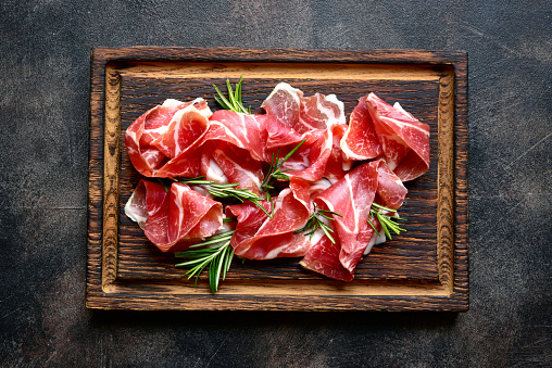 Slices of prosciutto di parma or jamon serrano (iberico) on a dark wooden background. Top view with copy space.