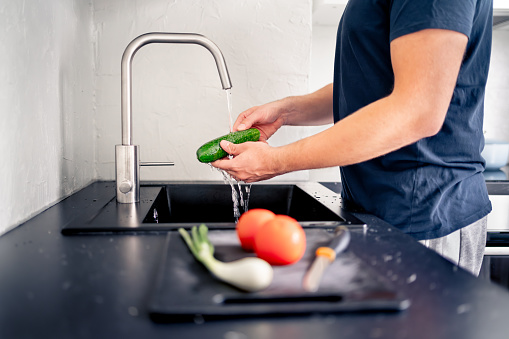 Washing vegetables in kitchen sink with water. Food preparation. Man cooking. Making salad. Cucumber in hand under faucet tap. Vegan or vegetarian meal. Home cook. Organic ingredients in recipe.