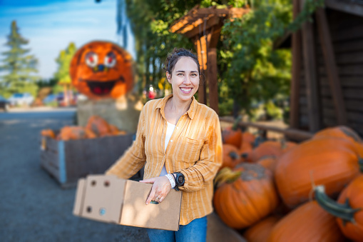 Portrait of an Eurasian woman standing next to a large crate of pumpkins smiling directly at the camera while holding a cardboard box filled with local produce. The woman is enjoying a day with her family at a harvest festival.