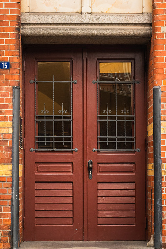 A vertical photograph captures a close-up view of a burnt-red door located at one of the historic buildings in Zollkanal, Germany. The door is framed by intricate ironwork on the windows, allowing glimpses into the interior. Flanking the door, part of the building's brick facade can be seen, with a decorative stone embellishment crowning the entrance. To one side of the door, the number 15 is clearly visible, providing a personal touch to this architectural detail. The image showcases the craftsmanship and unique character found in the urban landscape.