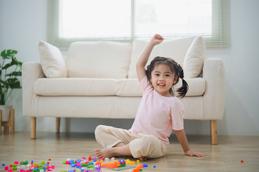 Joyful Asian girl happy and smiling raise her hand playing colorful plastic building block toys, sitting on the living room floor, creatively playing with plastic building block, building colorful structures creativity imagine.