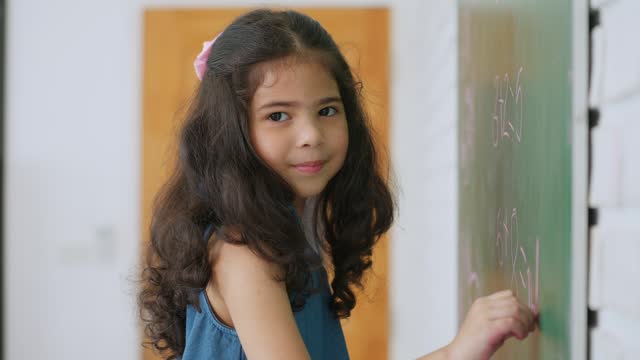 Asian little girl pupil writing or drawing on chalkboard smiling and looking at camera. Little schoolgirl smiling and looking at camera. Education and learning concept