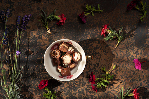 Handmade delicious chocolate candies of different shapes, from white and dark chocolate, in a porcelain cup, on a rustic brown background. Background is decorated with colorful flowers