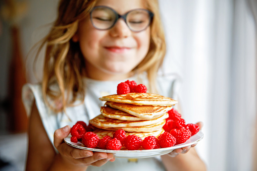 Little happy preschool girl with a large stack of pancakes and raspberries for breakfast. Positive child eating healthy homemade food in the morning