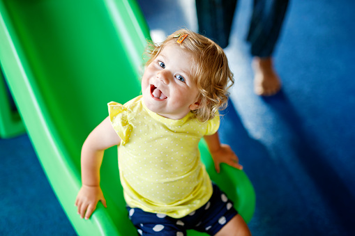 Happy blond little toddler girl having fun and sliding on indoor playground at daycare or nursery. Positive funny baby child smiling. Healthy girl climbing on slide