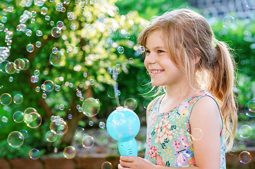 Happy little blonde preschool girl having fun with blowing soap bubble blower. Cute child playing on sunny summer day. Happy active funny healthy kid. Activity for children