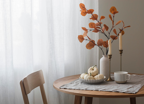 Autumn home interior in the living room - around wooden table, a chair, a bouquet of autumn leaves in a vase, decorative pumpkins. Cozy home