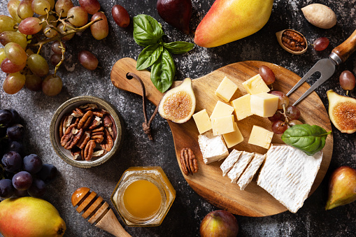 Brie cheese, fresh figs, grapes, cheese cubes, pears, pecan nuts and honey on cutting board on dark stone background. Cheese plate, still life of seasonal wine snacks. View from above.