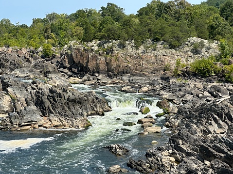 Cascades in Great Falls National Park, part of the George Washington Memorial Parkway, in McLean, Virginia. Water flowing over ancient rocks, tree area in the background and a clear blue summer sky overhead.