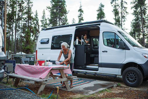 Active and healthy senior woman stands at campsite picnic table beside a sprinter van, preparing a meal with her granddaughter's help during a family vacation at an Oregon campground.