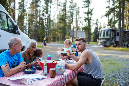 Active multi-racial senior couple sit with their multi-generation family at a picnic table in a campground and enjoy eating a meal outdoors while camping in the Pacific Northwest region of the United States.