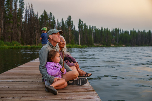 Active and healthy senior couple sit on a dock and cheerfully interact with their Eurasian preschool age granddaughter while watching the sun set over a lake during a multigenerational family camping trip.