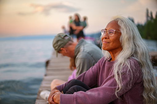 Side view of a beautiful and vibrant senior woman of Pacific Islander descent sitting on a dock with her multigenerational family and watching the sun set with a peaceful expression on her face on a warm summer evening in the Pacific Northwest region of the United States.