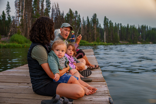 A cute Eurasian one year old baby boy sits in his mother's lap and looks directly at the camera while his multigenerational family enjoys a relaxing evening at the lake watching the sun set. The family is on a camping trip in Oregon.