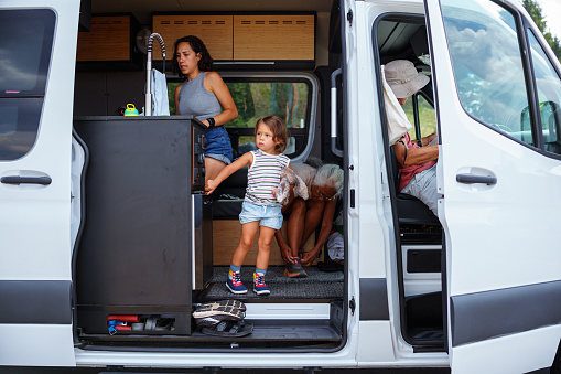 An Eurasian preschool age girl lovingly holds a stuffy toy and looks with curiosity out the open door of a camper van while on a camping trip in Oregon with her multi-generational family.