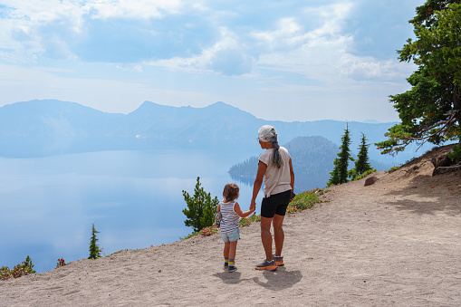 An active multiracial senior  woman of Hawaiian and Chinese descent holds hands with her three year old Eurasian granddaughter while hiking along a mountain ridge overlooking Crater Lake in Oregon on a summer day.
