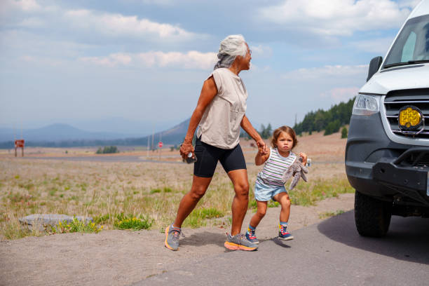 Grandchild with grandmother wearing a colorful summer dresses are walking along an alley in a tropical garden. Sunset sunlight bathes the landscape, with trees and lush foliage. Happy vacations.