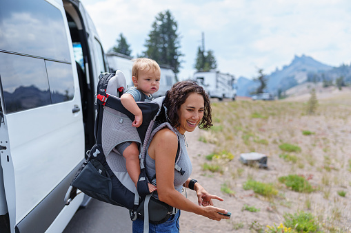 An active Eurasian woman stands outside a camper van and smiles while carrying her cute one year old baby boy in a backpack style baby carrier before going for a hike while on a camping trip in Oregon.