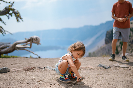 A cute three year old Eurasian girl squats in the dirt and draws with a stick while on a hike overlooking Crater Lake in Oregon while on a summer camping vacation with her family.