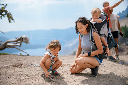 A cute three year old Eurasian girl who is on a hike with her family squats in the dirt and shows her mom a drawing she did with a stick. The multi-generation family is hiking along a trail overlooking Crater Lake in Oregon while on a summer camping vacation.