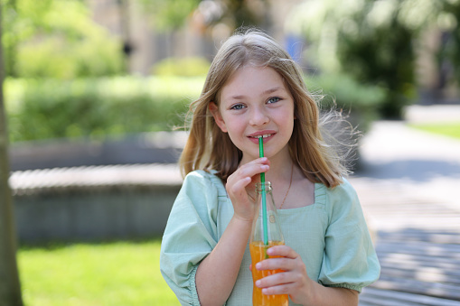 A cute young girl sipping a refreshing orange beverage through a straw in a park, enjoying a healthy and tasty treat.