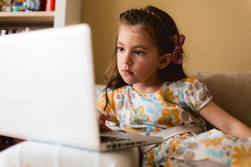 Little girl sitting on a sofa in the living room and watching on laptop