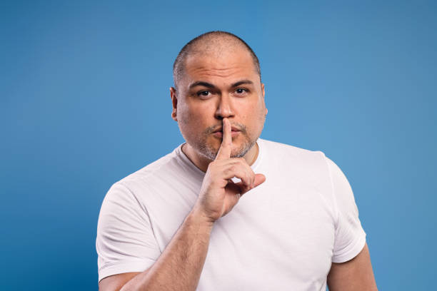 istock Portrait of a Brazilian man wearing a basic white shirt, facing forward, asking for silence, placing the index finger of his right hand close to his mouth, looking at the camera - Belém - Pará - Brazil 1668452738