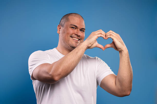 portrait of a brazilian man wearing a basic white shirt, from the front, making a heart with his hands, raised to his face, looking at the camera and smiling - belém - pará - brazil - human heart flash imagens e fotografias de stock