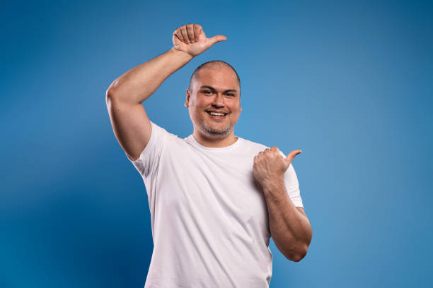istock Portrait of a Brazilian man wearing a basic white shirt, facing forward, pointing to the side with the thumb of both hands, smiling and looking at the camera - Belém - Pará - Brazil 1668452762