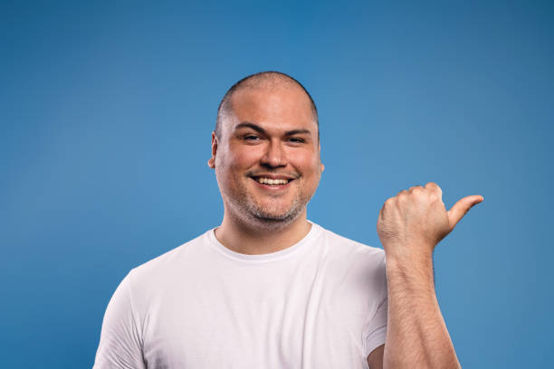 istock Portrait of a Brazilian man wearing a basic white shirt, facing forward, pointing to the side with the thumb of his left hand, smiling looking at the camera - Belém - Pará - Brazil 1668453566