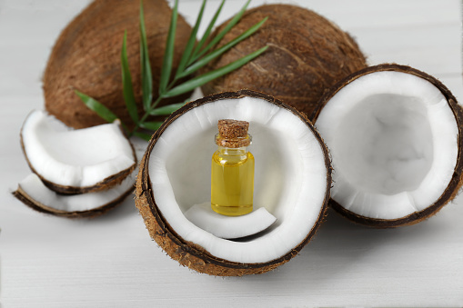 Bottle of organic coconut cooking oil, leaf and fresh fruits on white wooden table, closeup