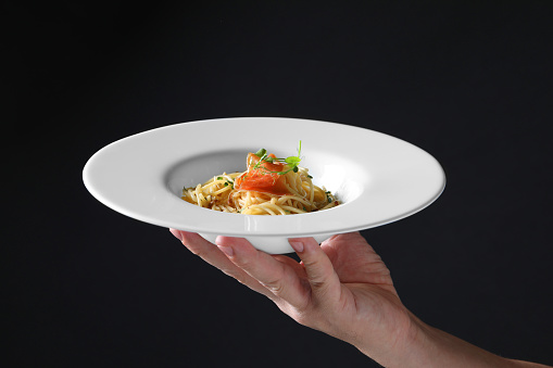 Woman holding plate of tasty spaghetti with prosciutto and microgreens on black background, closeup. Exquisite presentation of pasta dish