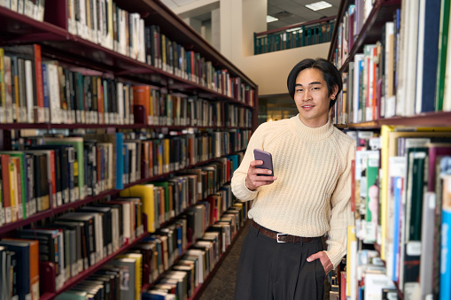 Portrait of smart Japanese university student holding mobile phone in bookstore copy space. Handsome asian man in modern library. Education concept