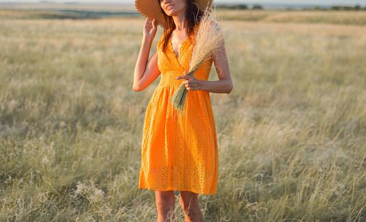 Young woman in an orange dress and a straw hat standing on a field in the rays of the setting sun.