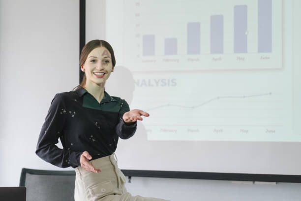 portrait of smart businesswoman standing at comfortable workplace and looking at somebody with calmness. joyful female pointing at charts and graphs. - conference room sign imagens e fotografias de stock