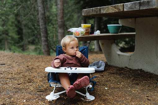 An adorable Eurasian one year old boy sits in a high chair next to a picnic table and eats while on a camping trip in Oregon with his family.