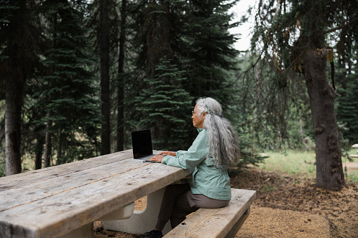 A vibrant senior woman of Pacific Islander descent who has found life balance by working remotely while traveling sits at a picnic table and uses a laptop computer to work while on a camping trip in the Pacific Northwest region of the United States.