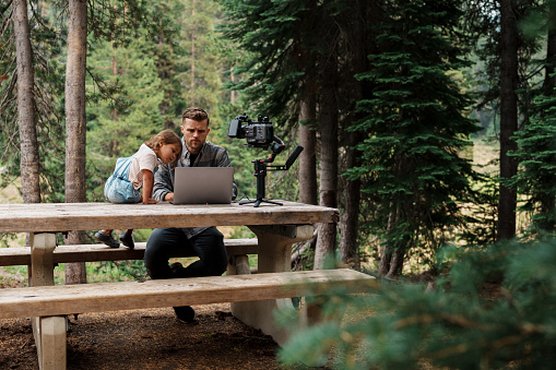 A man who works in media production sits at a campground picnic table and edits photos and videos as his curious three year old Eurasian daughter sits and watches. The man is working remotely while on vacation with his family.