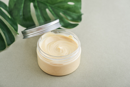 Open cosmetic cream container with green herbal leaves on wooden background closeup
