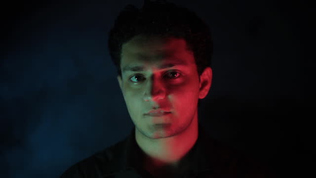 Close-up cinematic shot of a young male teenager in a black shirt sitting in a retro bar with neon lights falling on his face. Neo-noir light-up Hd stock footage. Man sitting in neon lights - 24 fps - ProRes