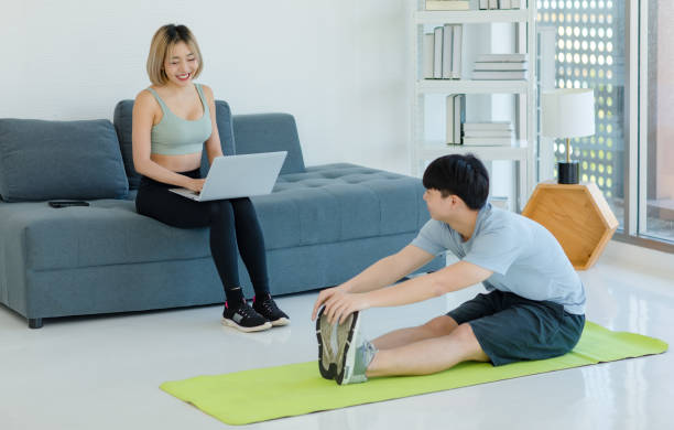 istock Asian male husband sitting on yoga mat body stretching warming up practicing training exercising yoga while young female wife in sportswear sport bra legging sneakers using laptop at home 1669185679