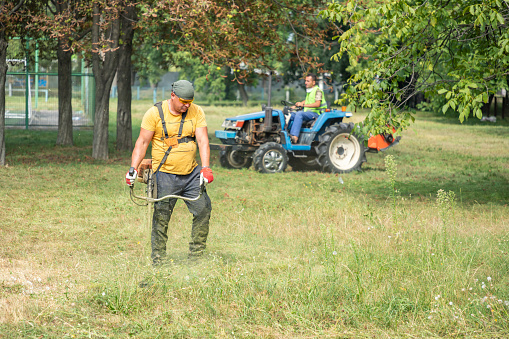 Man gardener mowing grass with electric or petrol lawn trimmer with male gardener on a Mini-tractor with a chain mower chopping, mows grass on a background. Maintenance of the territory, mulching of grass. Gardening care tools and equipment.