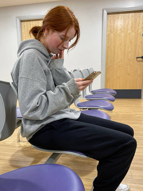 Stock photo showing a female patient waiting for an appointment in a doctor's waiting room, which is part of a hospital medical centre. The room has been designed to appear spacious and modern, being themed around the colour red, with seating that makes it easy to clean the floor beneath. imagen en primer plano de una atractiva mujer pelirroja en la sala de espera del médico en el hospital, sentada desplazándose en la pantalla del teléfono inteligente en una fila de asientos modernos y fáciles de limpiar, limpie los cojines de asiento c