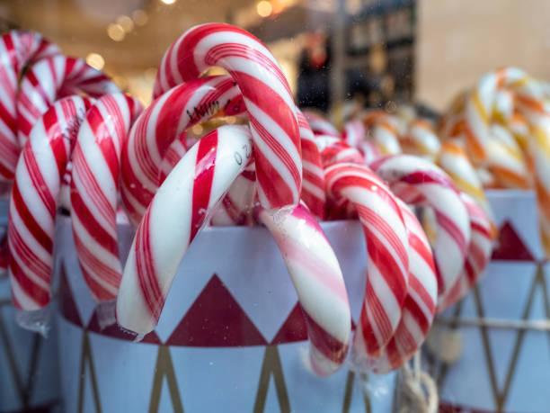 A vibrant red gift box featuring white cursive 'Merry Christmas' text and star accents sits prominently in the center of the image, surrounded by multiple red and white striped candy canes leaning against it. They are on a Santa hat with white fur fabric around the base, evoking snow or wrapping paper, creating a cosy holiday atmosphere. The composition highlights classic Christmas elements without any people present. This image captures the joyful essence of winter festivities, suitable for commercial uses such as holiday greeting cards, seasonal advertisements, e-commerce product displays for gifts and decorations, social media posts promoting Christmas sales, and editorial content in magazines or websites focused on holiday traditions and home décor. The bright red and white colour scheme emphasizes cheer and tradition, ideal for branding materials in retail, publishing, and media industries targeting winter holiday campaigns.