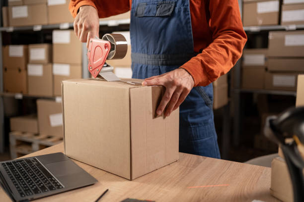 Worker man In Retail Warehouse working packing parcel box with tape dispenser for shipment. Delivery and distribution center full of shelves with goods. Close-up Worker man In Retail Warehouse working packing parcel box with tape dispenser for shipment. Delivery and distribution center full of shelves with goods. Close-up. Copy space logistical stock pictures, royalty-free photos & images
