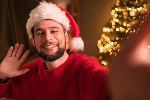 Happy bearded man in Santa Claus hat holding phone waving hand taking selfie, video calling, recording video Merry Christmas greeting or shooting vlog. Close-up