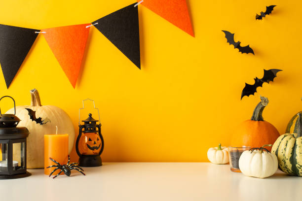 istock Spookify your table for Halloween. Side view image of a tabletop featuring themed decor, pumpkins, luminous jack-o'-lantern lamp, bats, spiders, candles, and festive flag garland on a yellow wall 1669269737